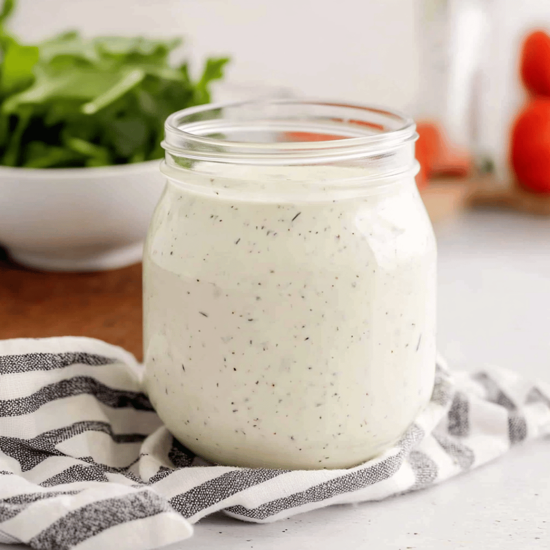 Creamy Homemade Hidden Valley Ranch Dressing in Glass Jar Glass jar of homemade Hidden Valley ranch dressing placed on a striped kitchen towel, with fresh greens and tomatoes in the background.