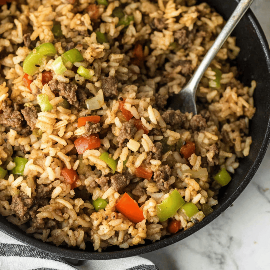 Dirty rice in a cast iron skillet with ground meat, green and red bell peppers, and a serving spoon.