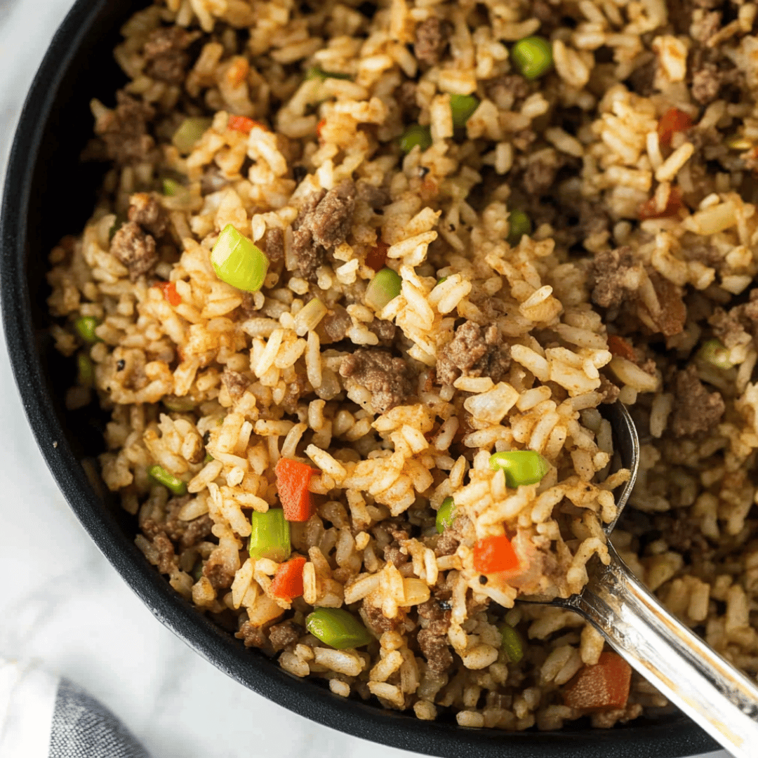 Classic Dirty Rice with Sausage and Peppers Close-up of dirty rice in a black bowl with ground meat, red and green bell peppers, and a metal spoon.