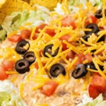 The Best Taco Dip Recipe: Easy, Layered, and Perfect for Parties 2 lose-up of creamy taco dip topped with shredded cheddar cheese, sliced black olives, diced tomatoes, and lettuce, served with tortilla chips.